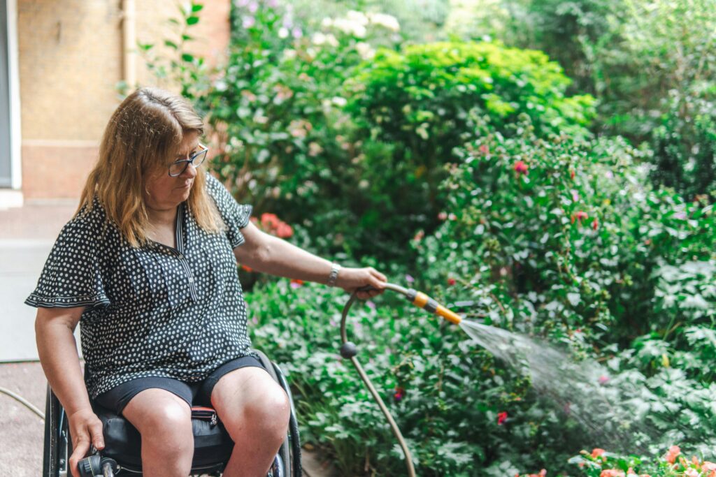 A woman in a wheelchair tending to her garden, watering plants with a hose.